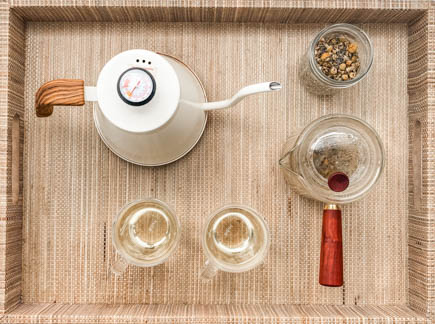 overhead view of wicker tray with white tea kettle, 2 glass mugs, clear teapot and dried chamomile and lavender flower buds