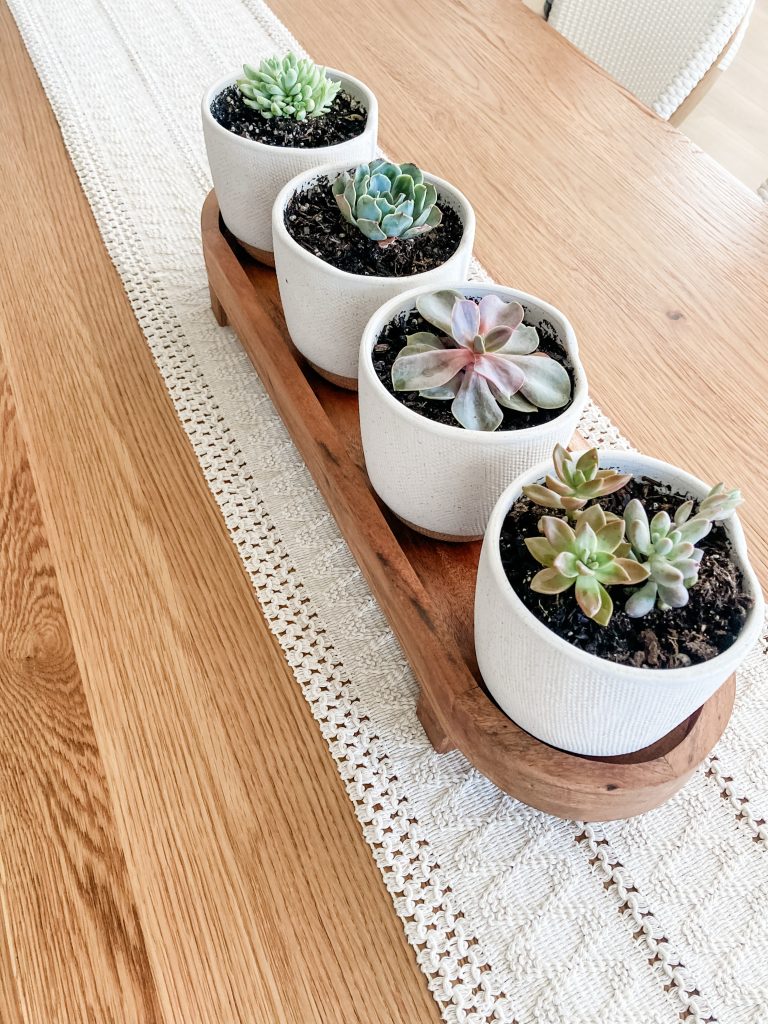 four small succulent plants in white pots on wooden tray on table runner displayed as centerpiece for table