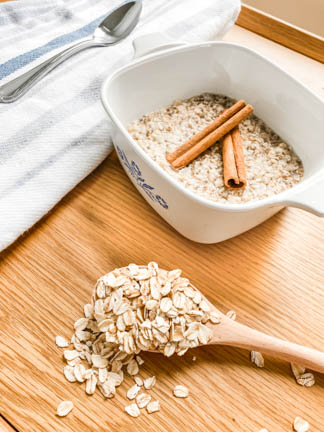 Oats spilling out of wooden spoon, white bowl with handles  containing warm oatmeal with cinnamon sticks on top.