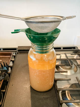Glass jar with beef bone broth showing funnel and strainer above it. Sitting on gas stove