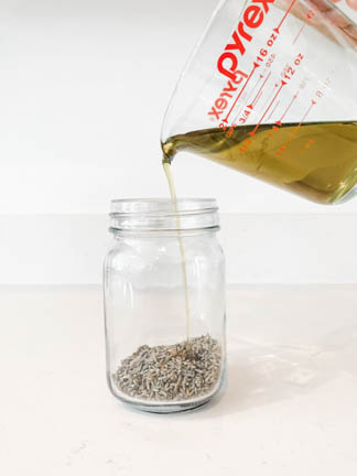 Glass jar holding dried lavender buds. Oil is pouring over lavender into jar.