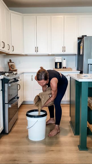 Woman pouring wheat berries into 5 gallon bucket in kitchen