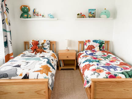 Child's bedroom featuring two twin beds with nightstand between them. Bedding and curtains are light colored dinosaur themed.