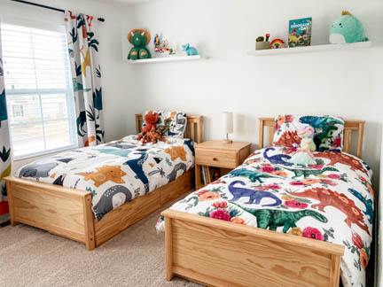 Child's bedroom featuring two twin beds with nightstand between them. Curtains hang from the window. Bedding and curtains are light colored dinosaur themed. 